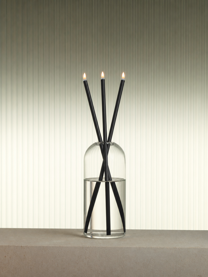 Azhara Eternal Candle Set: three lit black refillable oil candles in a clear glass vase on a stone plinth against a fluted wall